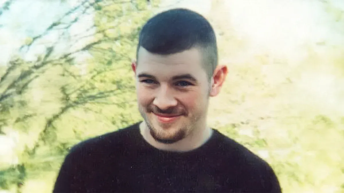 A smiling portrait of Eric Grant Cheeks, wearing a black shirt, with a blurred background of green foliage and sunlight. This image was commonly used in missing person flyers following his disappearance.