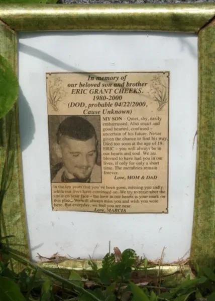 A framed memorial tribute to Eric Grant Cheeks, displaying his photo and messages from his parents and sister. The text highlights his family's love and enduring grief, expressing their longing and remembrance. The frame rests against his headstone, surrounded by greenery.