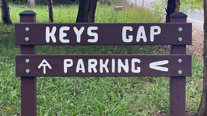 Wooden sign reading 'Keys Gap' with the Appalachian Trail symbol pointing straight ahead and 'Parking' with an arrow pointing left. The sign is located in a grassy area surrounded by trees, with a road visible in the background.