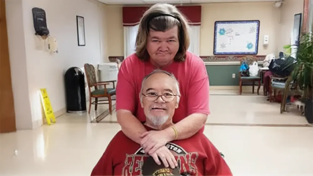 Richard and Joyce Lonas smiling together at a nursing home in 2018. Joyce stands behind Richard with her arms resting on his shoulders.