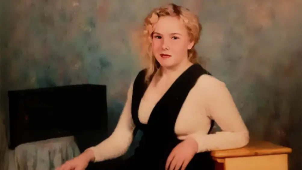 Susan Capino in a posed 1995 school portrait, seated with her hand on a yellow desk, wearing a black jumper over a cream top, in front of a mottled blue-gray studio backdrop.