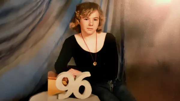 Portrait of Susan Capino taken in 1996. She wears a black top and moon necklace, seated beside a large "96" prop, against a soft blue and brown backdrop.